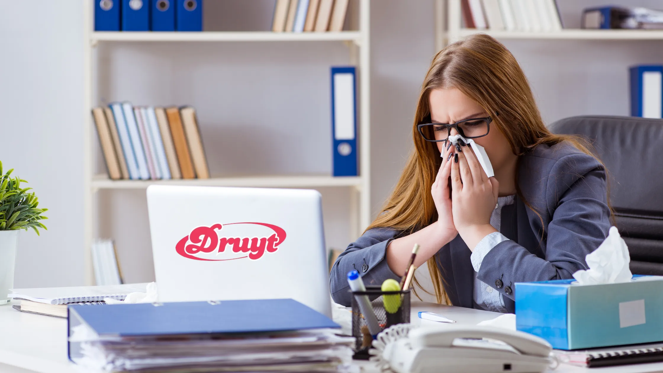 A woman in an office setting sneezes into a tissue, appearing unwell while sitting near an air conditioner. Focus keyword: "Can AC Cold Make You Sick?"