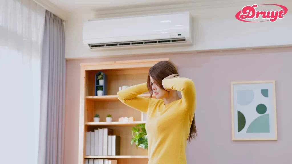 A woman in a room with an air conditioner holds her head, possibly feeling discomfort. Focus keyword: "Can AC Cold Make You Sick?"