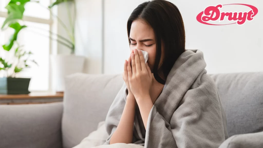 A woman wrapped in a blanket sneezes into a tissue, indicating she is feeling unwell. Focus keyword: "Can AC Cold Make You Sick?"